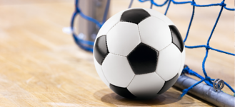 Piłka nożna leży w pobliżu linii bramkowej wewnątrz małej bramki na drewnianej podłodze, sugerując halową grę w piłkę nożną.