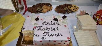 Na zdjęciu widać biały tort z napisem "Dzień Kobiet Im. Głusk" oraz wykrojonym kawałkiem, otoczony talerzami z różnymi ciastkami i słodyczami.