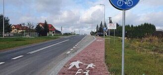 Droga z pasem ruchu dla rowerów po prawej stronie i chodnikiem, znak drogowy wskazujący ścieżkę rowerową, domy i niebo z chmurami w tle.
