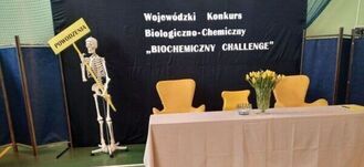 Na zdjęciu widoczny jest sztuczny szkielet obok banera z napisem "Wojewódzki Konkurs Biologiczno-Chemiczny BIOCHEMICO CHALLENGE" oraz stół z trzema pomarańczowymi krzesłami i bukietem żółtych tulipanów.