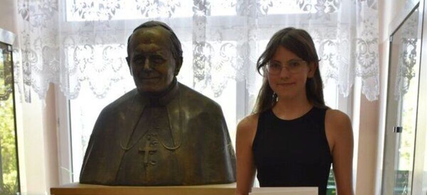 Młoda kobieta stoi obok biustu osoby, trzymając dokument. Biust jest umieszczony na postumencie z napisem "JAN PAWEŁ II 1920-2005". Za oknem widoczne drzewa.