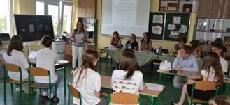 Klasa szkolna z uczniami siedzącymi przy ławkach oraz nauczycielką stojącą z przodu, tablica z prezentacją w tle.