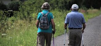 Starsza para spaceruje po ścieżce w parku, używając kijków nordic walking. Noszą wygodne ubrania na letni dzień.