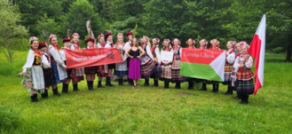Grupa osób w tradycyjnych polskich strojach ludowych trzyma banery i flagę Polski na tle zielonej łąki i drzew.