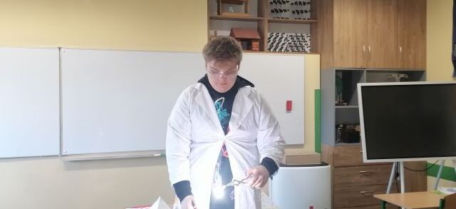 Uczeń w białym fartuchu stoi w klasie, trzymając lampę nad stołem. W tle widoczne są tablica, zegar, szafka z modelami cząsteczek, plakat kosmiczny i ławki szkolne.