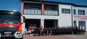 Grupa strażaków w galowych mundurach stoi na placu przed nowoczesną strażnicą. Obok widoczne są dwa wozy strażackie. Na budynku napis „Ochotnicza Straż Pożarna”. Na środku osoba trzyma sztandar.