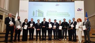 Grupa osób w formalnych strojach stoi w rzędzie, trzymając dokumenty, na tle ekranu z napisem konferencji "Renowacja budynków". W tle widać dużą salę z organami.