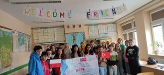 Grupa dzieci i dorosłych pozuje w sali lekcyjnej z flagami Polski i Turcji. Nad nimi widnieje napis "Welcome Friends". W tle mapy i plansze edukacyjne.