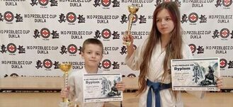 Chłopiec i dziewczynka w strojach do karate pozują z dyplomami i pucharami na tle baneru turniejowego.