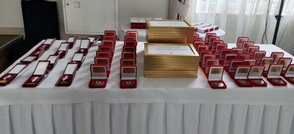Na zdjęciu znajduje się stół przykryty białym obrusem, na którym ułożone są liczne medale w czerwonych pudełkach oraz stosy złotych certyfikatów oprawionych w ramki. W tle widoczne jest okno i zasłona.