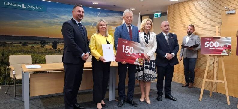 Pięć osób stoi w sali konferencyjnej. Dwie osoby trzymają duże czeki. W tle widoczny jest pejzaż z napisem "lubelskie".