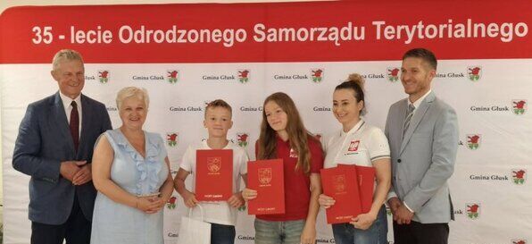 Grupa osób pozuje na tle baneru z napisem o 35-leciu Odrodzonego Samorządu Terytorialnego. Dwoje dzieci trzyma dyplomy. Obok stoją dorośli, dwie kobiety i dwóch mężczyzn, wszyscy ubrani elegancko.