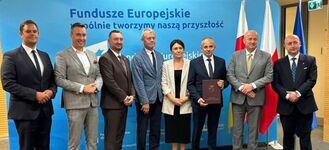Grupa ośmiu elegancko ubranych osób stoi na tle niebieskiego banera z napisem "Fundusze Europejskie". Na banerze widać także polskie i unijne flagi. Jedna osoba trzyma dokument.