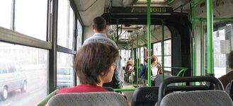 Wnętrze autobusu, widok na rząd pustych siedzeń oraz kilku pasażerów siedzących z przodu. Pasażerka z krótkimi włosami siedzi tyłem do fotografa. Przez okno widoczna jest ulica.