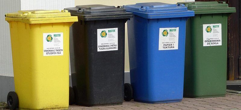 Cztery kolorowe pojemniki do segregacji odpadów stoją na chodniku przed budynkiem. Żółty, czarny, niebieski i zielony kosz są przygotowane na różne rodzaje śmieci.