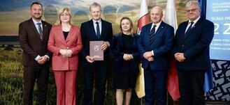 Grupa sześciu osób stoi na tle panelu z napisami i flagami. Jedna z osób trzyma teczkę, reszta uśmiecha się do kamery. W tle widnieje zdjęcie krajobrazu.