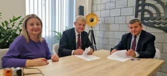 Trzy osoby siedzą przy stole i podpisują dokumenty. Na stole leżą papiery i długopisy. Za nimi jest lampa stojąca i ściana z dekoracyjnym wzorem. W tle widać rośliny i żaluzje.