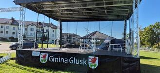 Scena plenerowa w słoneczny dzień, przygotowana na wydarzenie w gminie Głusk. Na scenie znajdują się puste krzesła. W tle widoczne są budynki mieszkalne i drzewa.