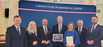 Na zdjęciu siedem osób stoi przed niebieską ścianką z napisem "Lubelski Urząd Wojewódzki w Lublinie". Dwoje z nich trzyma certyfikaty. Grupa jest elegancko ubrana, cztery osoby to mężczyźni, trzy to kobiety.