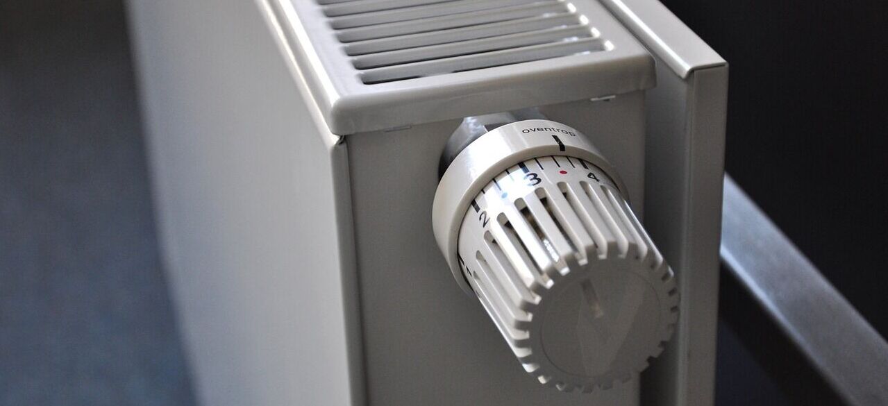 Zdjęcie przedstawia biały kaloryfer z widocznym pokrętłem termostatycznym ustawionym na poziom 3. Kaloryfer stoi na tle ciemnej ściany, a podłoga jest pokryta niebieską wykładziną.