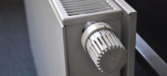 Zdjęcie przedstawia biały kaloryfer z widocznym pokrętłem termostatycznym ustawionym na poziom 3. Kaloryfer stoi na tle ciemnej ściany, a podłoga jest pokryta niebieską wykładziną.