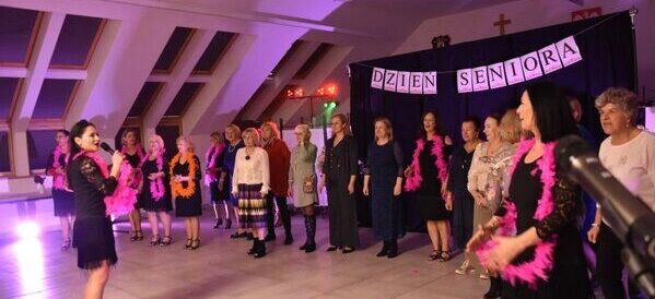 Grupa osób starszych, głównie kobiet, tańczy w sali ozdobionej na Dzień Seniora. Uczestnicy noszą kolorowe boa i stoją w półkolu, patrząc na występ. Sala ma ozdobne światła i napis „Dzień Seniora”.