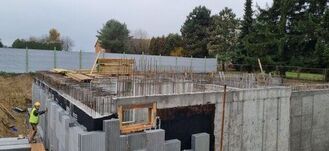 Na zdjęciu widoczna jest budowa z betonowymi ścianami fundamentowymi. Na pierwszym planie widać pracownika w kamizelce odblaskowej i kasku. W tle znajdują się drzewa i ogrodzenie.