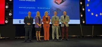 Pięć osób stoi na scenie przed ekranem z napisem "Fundusze Europejskie" i flagą UE. Osoby są elegancko ubrane, a obok nich znajdują się flagi Polski i UE.