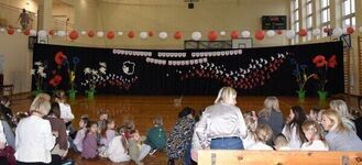 Sala gimnastyczna udekorowana biało-czerwonymi flagami i plakatami. Na środku sceny motywy kwiatowe i godło Polski. Dzieci i dorośli siedzą na podłodze skupieni na wydarzeniu.