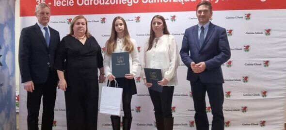 Pięć osób stoi na tle banera z napisem "35-lecie Odrodzonego Samorządu Terytorialnego". Dwie osoby trzymają dyplomy i torby. Wszyscy mają formalne stroje. W górnym rogu wisi godło Polski.