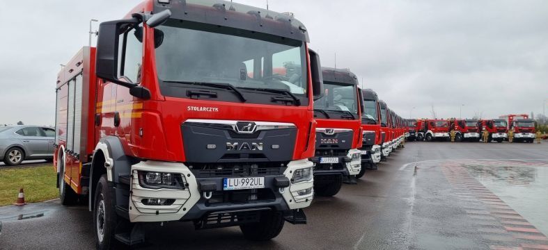 Strażackie pojazdy ustawione w szeregu na parkingu. Każdy z nich jest czerwony z charakterystycznymi oznaczeniami, gotowy do wyruszenia na akcję. Pogoda pochmurna, asfalt mokry od deszczu.