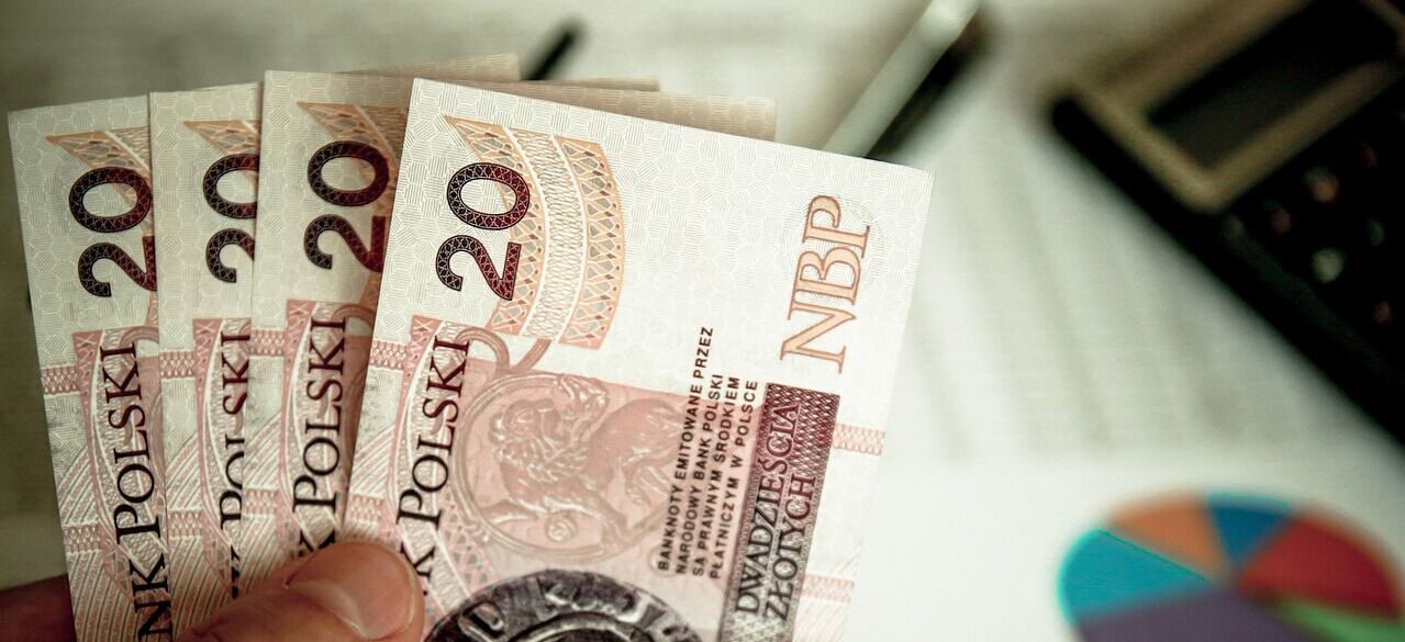 Osoba trzyma w ręku kilka banknotów o nominale 20 złotych. W tle znajduje się wykres kołowy i kalkulator, sugerujące kontekst finansowy lub biurowy.