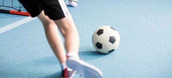 Osoba ubrana w czarne spodenki kopie piłkę nożną na niebieskim boisku wewnętrznym. W tle widać fragment bramki i drugą osobę. Atmosfera sugeruje rekreacyjną grę w halową piłkę nożną.