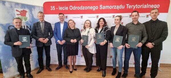 Grupa osób pozuje na tle baneru z napisem "35-lecie Odrodzonego Samorządu Terytorialnego". Każda osoba trzyma teczkę z herbem. W tle widoczna jest dekoracja z logo gminy.