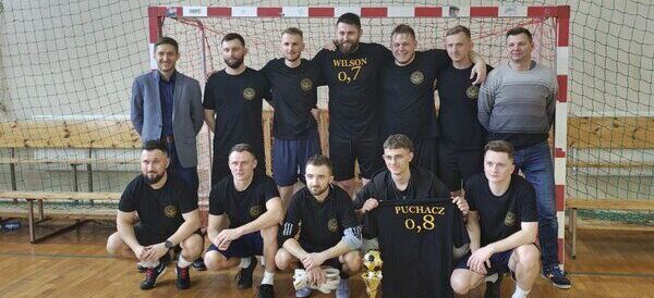 Grupa mężczyzn w czarnych koszulkach piłkarskich pozuje z pucharem na tle bramki do piłki nożnej w hali sportowej. Trzech mężczyzn stoi w dwóch rzędach, a za nimi jest siatka.