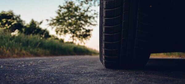 Zbliżenie opony samochodowej na asfaltowej drodze. W tle widać zarys rozmytego drzewa i zieloną trawę przy drodze oświetloną ciepłym światłem zachodzącego słońca.