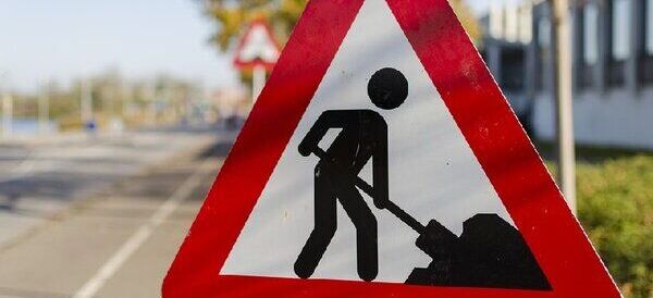 Znak ostrzegawczy w kształcie trójkąta z czerwonym obramowaniem przedstawia sylwetkę osoby pracującej nad kopaniem ziemi. W tle rozmyta droga i drzewa.