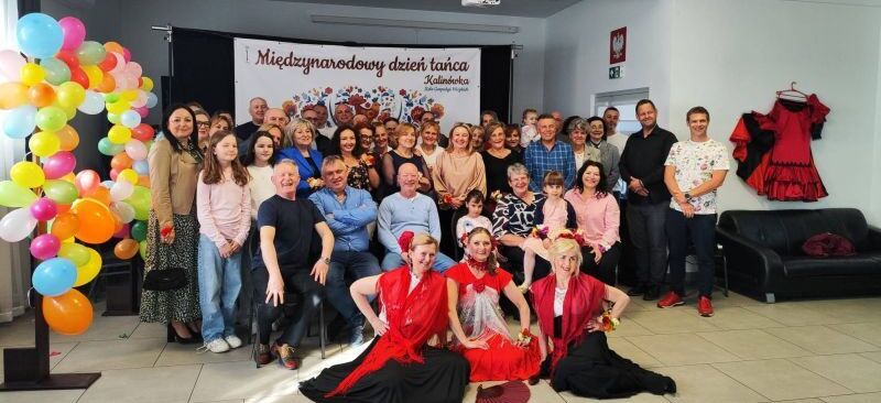 Grupa ludzi pozuje do zdjęcia w sali ozdobionej balonami i kwiatami z okazji Międzynarodowego Dnia Tańca. Na pierwszym planie kobiety w hiszpańskich strojach siedzą na podłodze.