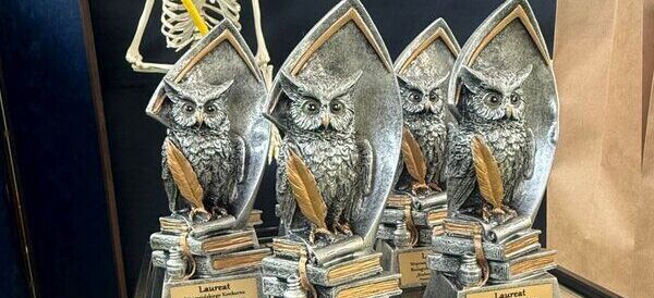 Kilka statuetek w kształcie sowy z piórem na podstawce. Obok figurki szkieletu człowieka i baner z napisem "POWODZENIA!". To scena z konkursu biologiczno-chemicznego.