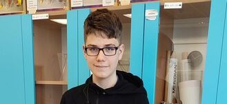 Uczeń w okularach stoi przed szafkami w klasie matematycznej. Za nim widać półki z bryłami geometrycznymi i planszami edukacyjnymi. Ma na sobie czarną bluzę, a wnętrze klasy jest dobrze oświetlone.