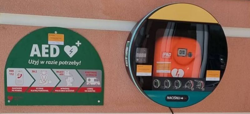 Na ścianie znajduje się koło zewnętrzne z automatycznym defibrylatorem (AED). Obok jest zielona tablica z instrukcjami użycia AED w nagłych przypadkach.