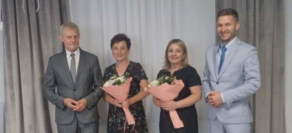 Cztery osoby stoją na tle zasłon. Dwie kobiety w środku trzymają bukiety kwiatów. Wszyscy są elegancko ubrani w formalne stroje.