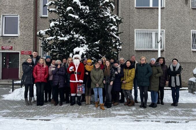 Grupa ludzi pozuje na zewnątrz w zimowej scenerii obok Świętego Mikołaja, w tle budynek i drzewo pokryte śniegiem.