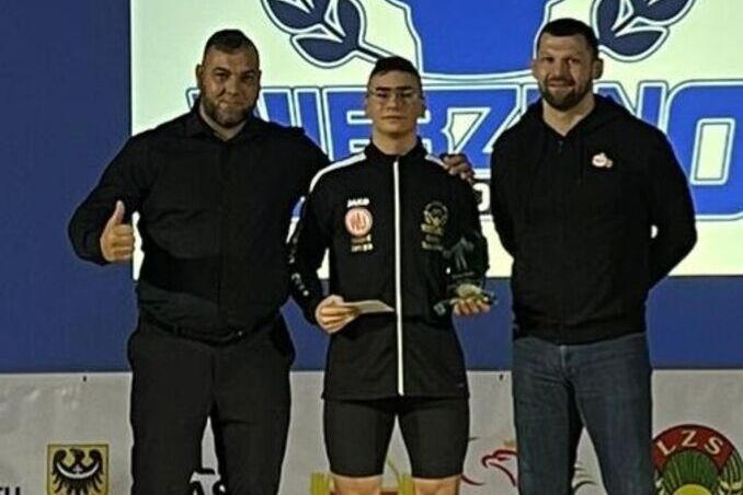 Trzy osoby stoją na podium sportowym, osoba po środku trzyma medal i puchar, po obu stronach stoją mężczyźni w sportowych ubraniach, wszyscy uśmiechnięci.