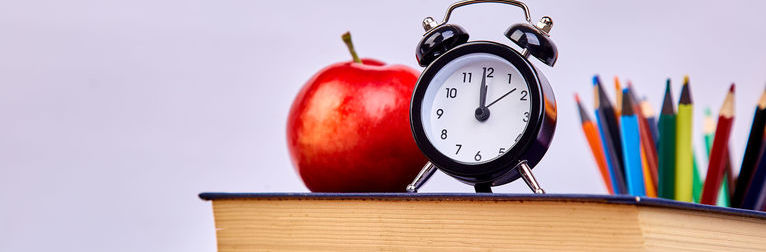 Grafika przedstawia kredki oraz książki na których stoi jabłko i zegar