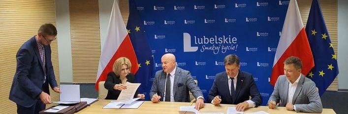 4 osoby siedzące przy biurku, jedna osoba stojącą obok na tle ścianki lubelskie i 2 flag polski oraz 1 unijnej