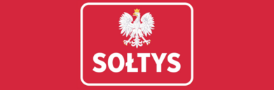 Czerwony baner z białą obwódką, na środku biały orzeł w koronie, pod nim napis "SOŁTYS" białymi literami na czerwonym tle.