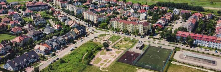 Zdjęcie lotnicze przedmieścia z mieszkanymi blokami, domami jednorodzinnymi, zielenią, boiskiem i ulicami.