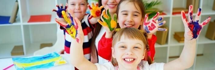 Grupa uśmiechniętych dzieci pokazuje dłonie pomalowane farbami, w tle widoczne są półki z przyborami szkolnymi.