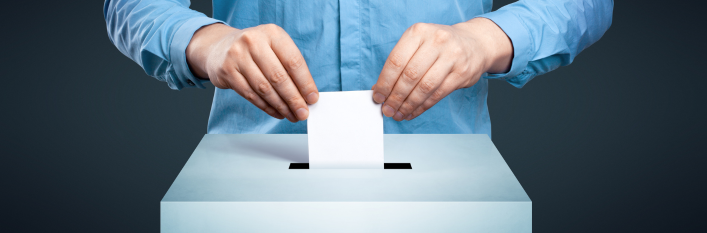 Osoba w niebieskiej koszuli wrzuca kartkę do głosowania do białej urny na ciemnoniebieskim tle.